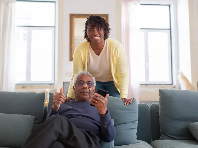 A younger woman supports an older relative as he uses a smartphone at home.